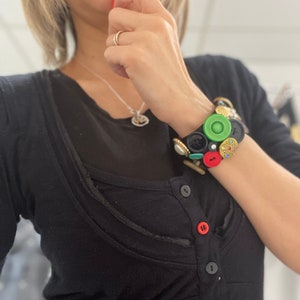 Puede incluir: Una camisa negra de manga larga con botones en la parte delantera. La camisa tiene un brazalete de botones decorativo en la muñeca, con una variedad de botones coloridos, incluyendo rojos, verdes y dorados.