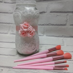 May include: A set of six pink makeup brushes with rose gold accents. The brushes are arranged in a fan shape on a white surface. A glass jar decorated with white lace and pink roses is in the background.