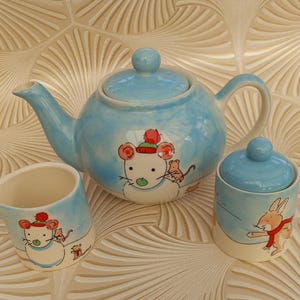 Puede incluir: Un juego de té de cerámica azul claro con tetera, jarra de crema y azucarero. Cada pieza está decorada con una ilustración caprichosa de un ratón con sombrero y un conejo con bufanda roja. El juego está diseñado para servir té.