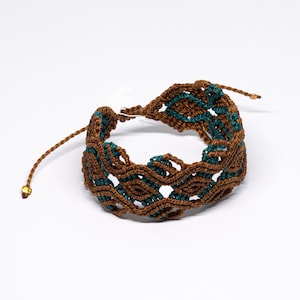 Peut inclure: Bracelet macramé fait main, marron et turquoise. Le bracelet présente un motif tissé avec des nœuds complexes, créant un aspect texturé. Le bracelet est réglable et possède une petite perle dorée au bout du cordon.