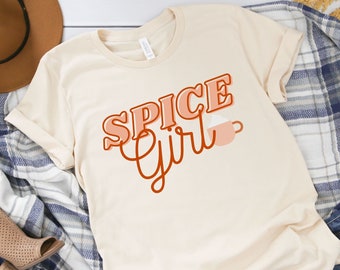 May include: A beige t-shirt with the text "Spice Girl" in orange and red lettering with a stylized coffee cup graphic. The shirt is laid out on a white surface with a plaid blanket, a brown hat, and blue jeans.