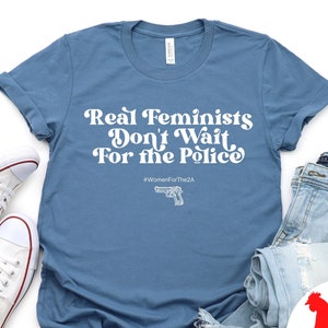 May include: A blue t-shirt with the text "Real Feminists Don't Wait For the Police" in white. The shirt also has the hashtag "#WomenForThe2A" and a silhouette of a handgun.