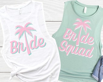 May include: Two white tank tops with pink palm trees and the text "Bride" and "Bride Squad" on them. The tank tops are on a white wooden background.