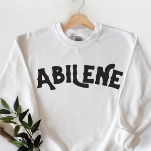 May include: White crewneck sweatshirt with the word "ABILENE" in large, black, stylized letters across the chest. The sweatshirt is hanging on a wooden hanger. A sprig of greenery is placed on the lower left side.