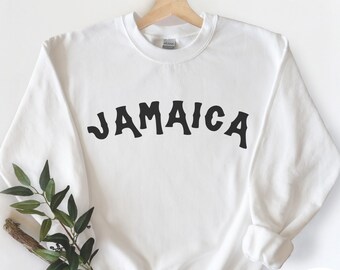May include: White sweatshirt with the word "JAMAICA" in bold, black, arched lettering. The sweatshirt is displayed on a wooden hanger. A sprig of greenery rests in front of the sweatshirt.