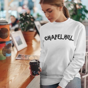 May include: A white sweatshirt with the text "CHAPEL HILL" printed in black on the front. The sweatshirt is being worn by a person holding a black and white floral patterned coffee cup.