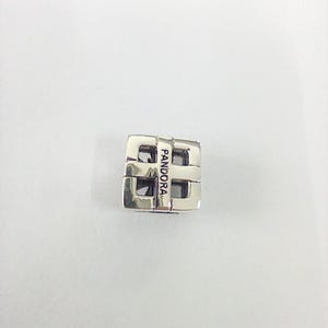 May include: A silver Pandora charm with a geometric design. The charm is a cube shape with open spaces and the word "PANDORA" engraved on one side. The charm is made of a shiny, reflective metal.