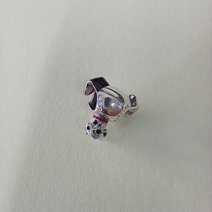 Puede incluir: Un charm de plata con forma de perro de dibujos animados, con orejas negras, un collar rojo y manchas negras en las patas. El charm tiene las palabras "©Disney" y "PANDORA" grabadas.