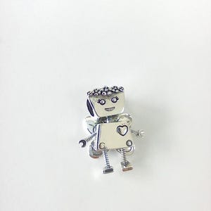 Puede incluir: Broche plateado con forma de robot y detalle de corazón. El robot tiene una cara sonriente, una corona y brazos y piernas pequeños. El broche está sobre un fondo blanco. Una joya encantadora.