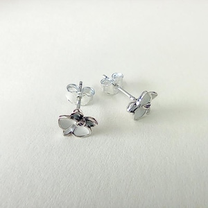 May include: A pair of silver-toned stud earrings. Each earring features a small, white flower design with delicate petals and a dark center. The earrings are displayed on a white surface, showcasing their intricate details and elegant style.
