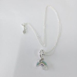 Puede incluir: Un collar de plata con un colgante de arcoíris. El colgante presenta un diseño de arcoíris colorido con una nube blanca. El collar está sobre un fondo blanco. Es una joya delicada.