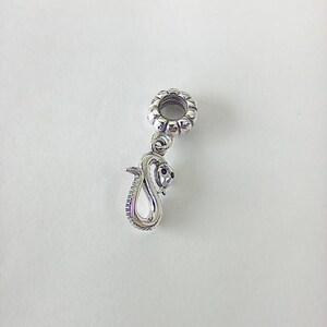 May include: A silver snake charm. The charm depicts a coiled snake with a detailed head and black eyes. The charm has a decorative top with a floral pattern, suitable for a bracelet or necklace. The charm is made of silver.