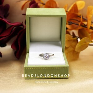 May include: A silver ring with a large, round, clear stone surrounded by smaller stones. The ring is in a green velvet ring box with the words "BeadsLondonShop Handcrafted Jewellery" on the bottom.