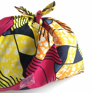 May include: A colorful fabric gift wrap with a pink, yellow, orange, and blue pattern. The fabric is tied with a knot at the top and has a small pink label with the letters "MP" on it.