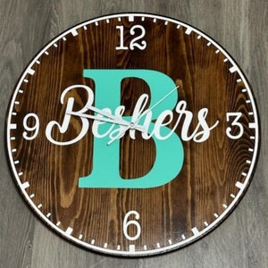 Può includere: Un orologio in legno con una lettera B turchese e il nome "Beshers" in scrittura bianca. Il quadrante dell'orologio ha numeri e lancette bianchi.