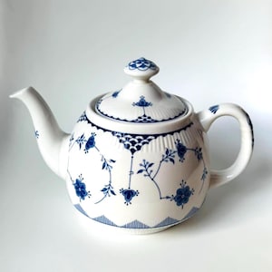 Puede incluir: Una tetera de porcelana blanca con un delicado patrón floral azul y detalles festoneados. La tetera incluye una tapa a juego con un pequeño pomo, un pico curvo y un asa resistente, todo ello decorado con motivos de color azul.