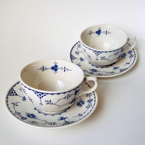 Puede incluir: Dos tazas de té de porcelana blanca con platos a juego que presentan un delicado patrón floral azul. Cada taza tiene un asa curva y un borde festoneado, mientras que los platos muestran una textura acanalada y motivos botánicos azules.