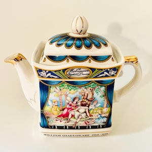 May include: A white ceramic teapot with a lid, featuring a scene from "A Midsummer Night's Dream." The teapot has gold accents and a blue and white floral design on the lid. The base of the teapot has the text "William Shakespeare 1564-1616."