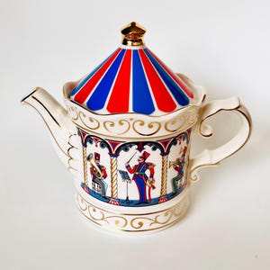 May include: A white ceramic teapot featuring a carousel-style lid with red and blue stripes and a gold finial. The body is decorated with gold scrollwork and illustrations of a marching band in blue and red uniforms under arched columns.