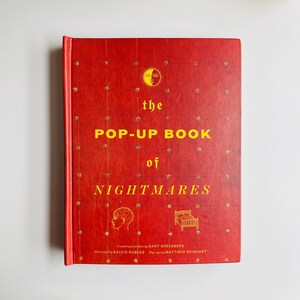 May include: A red hardcover book titled The Pop-Up Book of Nightmares with yellow text and gold star accents. The cover features a sun icon, a line drawing of a human head, and a bed. It lists Gary Greenberg, Balvis Rubess, and Matthew Reinhart.