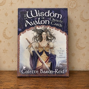 以下が含まれることがあります： コレット・バロン＝リードによる「The Wisdom of Avalon Oracle Cards」デッキとガイドブック。箱には52枚のカードデッキが含まれており、紫と白の背景に、長い黒髪で大きな剣を持った人物のイラストが描かれています。