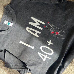 Puede incluir: Una camiseta gris con texto blanco que dice "I AM 40+" y un signo de paz blanco con esmalte de uñas rojo.