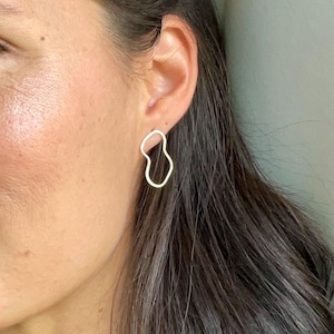 May include: A close-up of a person wearing gold-colored, abstract-shaped earrings. The earrings have an open, irregular design. The person has long, dark hair and is looking towards the left side of the frame. The background is a light, neutral color.