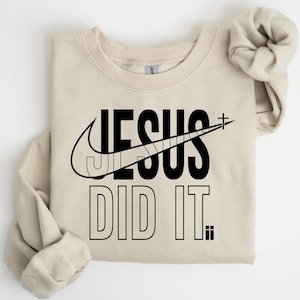 A beige t-shirt with a graphic design featuring the text "JESUS DID IT" and a Nike swoosh logo.