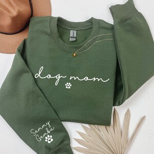 A green sweatshirt with the text "dog mom" printed on it, along with paw prints and a necklace. The sweatshirt is placed on a white surface, with a brown hat and a palm frond nearby.