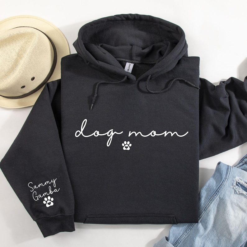 A black hoodie with the text "dog mom" printed on it, along with paw prints, is displayed alongside a hat and some clothing items.