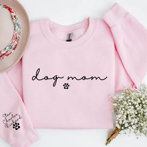 A pink sweatshirt with the text "dog mom" printed on it, along with paw prints, is displayed alongside a hat, a bouquet of white flowers, and a small plant.