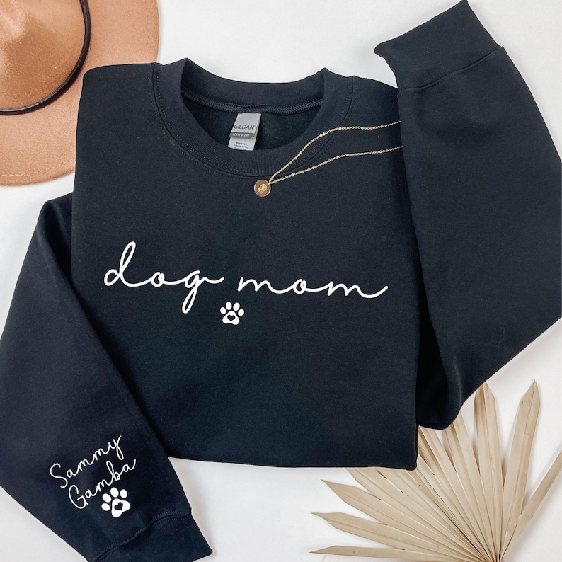 A black sweatshirt with the text "dog mom" printed on it, along with paw prints and a necklace. The sweatshirt is laid out on a white background, with a hat and some leaves visible in the background.