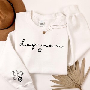 A white sweatshirt with the text "dog mom" printed on it, along with paw prints, is displayed alongside a brown hat and a palm frond.
