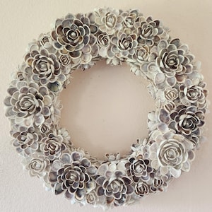 May include: A circular wreath constructed from numerous small, layered seashells arranged to resemble blooming flowers. The shells feature natural shades of white, cream, and light brown, with small white beads centered in several of the shell clusters.