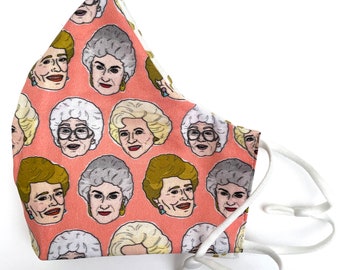 May include: A face mask in a salmon pink color featuring a repeating pattern of illustrated portraits of four different women with various hairstyles, including blonde and grey hair, some wearing glasses and earrings, with white elastic ear loops.