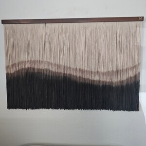 May include: A wall hanging made of yarn with a gradient of brown, beige, and black colors. The yarn is woven into a pattern of horizontal lines, creating a wave-like effect. The wall hanging is mounted on a wooden frame.