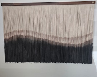 May include: A wall hanging made of yarn with a gradient of brown, beige, and black colors. The yarn is woven into a pattern of horizontal lines, creating a wave-like effect. The wall hanging is mounted on a wooden frame.