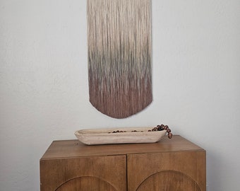 May include: A wall hanging featuring a dark wood top bar with a small circular logo, suspending numerous vertical threads that transition in color from a light cream at the top to a deep brown at the bottom in a rounded, ombre-style pattern.