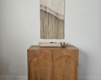 May include: A wooden cabinet with two doors, each with an arched top. The cabinet is a light brown color and has a wood grain pattern. A wooden tray with a beaded necklace sits on top of the cabinet.