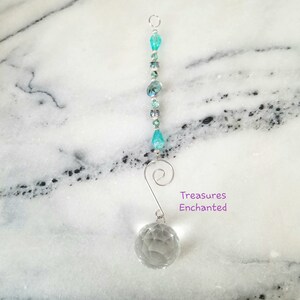 Peut inclure: Un attrape-soleil en cristal avec une boule de cristal transparente et un fil d'argent avec des perles turquoise. L'attrape-soleil a un crochet argenté en haut et le texte "Treasures Enchanted" est imprimé sous la boule de cristal.