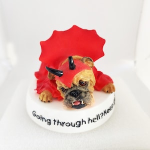 Könnte beinhalten: Eine kleine Figur eines Bullterriers, der als Teufel verkleidet ist und ein rotes Kostüm mit schwarzen Hörnern trägt. Die Figur sitzt auf einem weißen Sockel mit dem Text "Going through hell? Keep going!"