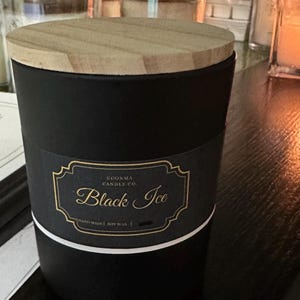 May include: A black candle with a wooden lid. The candle has a label that reads "Black Ice" in gold script. The label is on a black background with a gold border and white stripe.
