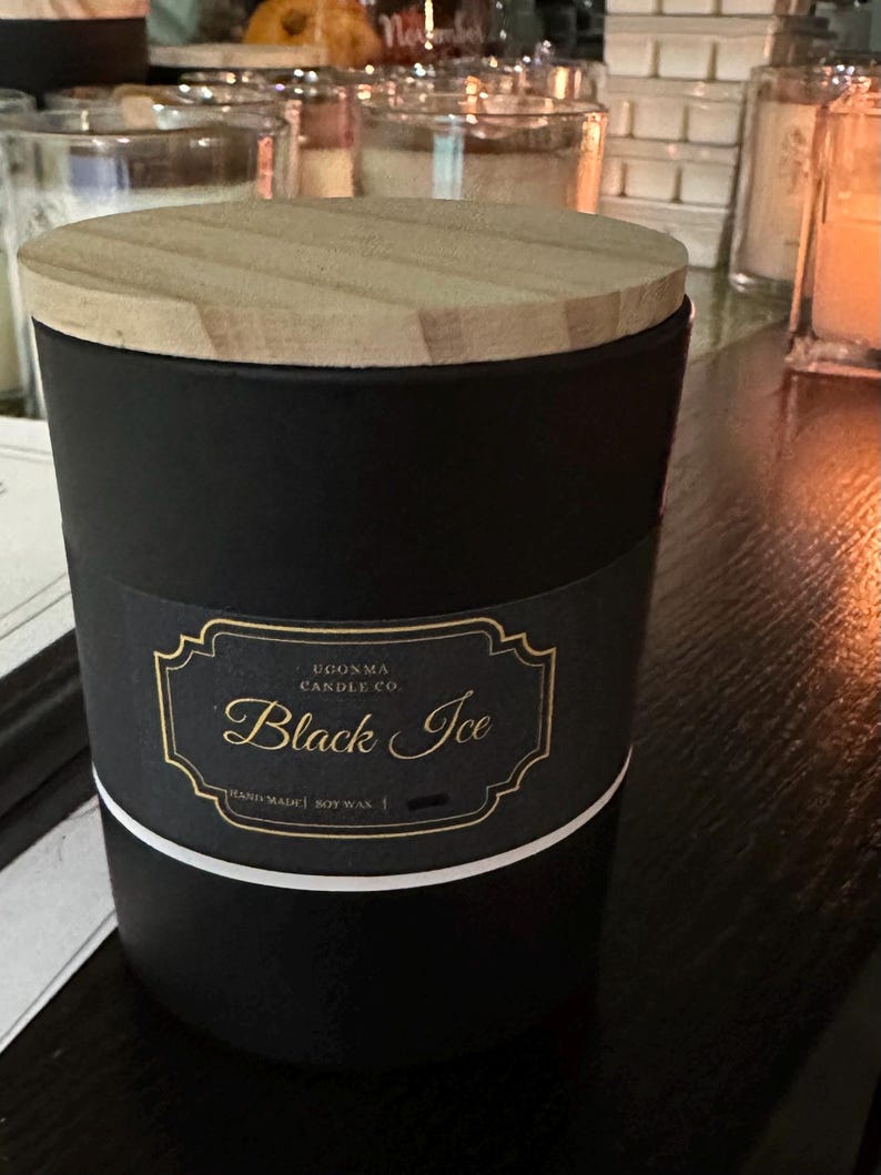 May include: A black candle with a wooden lid. The candle has a label that reads "Black Ice" in gold script. The label is on a black background with a gold border and white stripe.