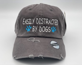 May include: A gray distressed baseball cap with the text "Easily Distracted By Dogs" embroidered in white with blue paw prints.
