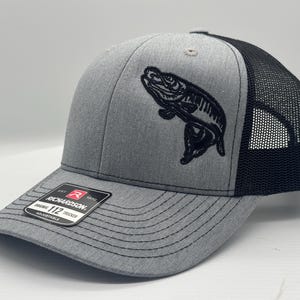 May include: A gray and black trucker hat with a black embroidered fish design. The hat has a black mesh back and a gray front with a black Richardson 112 label.
