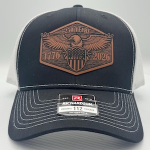 May include: Black and white trucker hat with a brown leather patch. The patch features an eagle with outstretched wings, the text "250 YEARS 1776-2026 MADE IN AMERICA", and a shield with stripes. The hat is adjustable.
