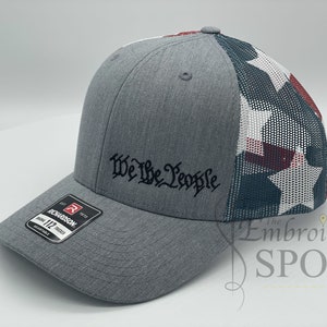 May include: A gray and white baseball cap with a mesh back. The front of the cap has the words "We The People" embroidered in black. The back of the cap has a red, white, and blue star pattern.