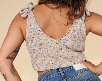 May include: A cream-colored knit top with blue flecks and tie straps. The V-neck tank top is paired with blue denim jeans. The top has a ribbed texture and a cropped fit, offering a casual and stylish look.