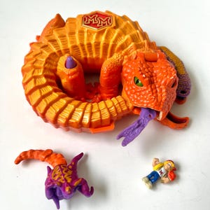 Puede incluir: Una serpiente de juguete naranja y amarilla enrollada, con una lengua púrpura, ojos verdes y las letras "MM" en la espalda. Incluye una criatura con cuernos morada y naranja y una pequeña figura de acción.