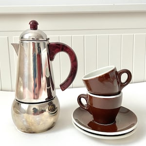 Peut inclure: Une cafetière italienne en acier inoxydable avec une poignée et un couvercle bordeaux. Deux tasses à expresso et soucoupes marron sont empilées à proximité. Les tasses ont des intérieurs blancs et les soucoupes ont un bord blanc.